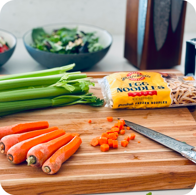 A cutting board with chicken noodle soup ingredients and a package of Grandma's Frozen Egg Noodles