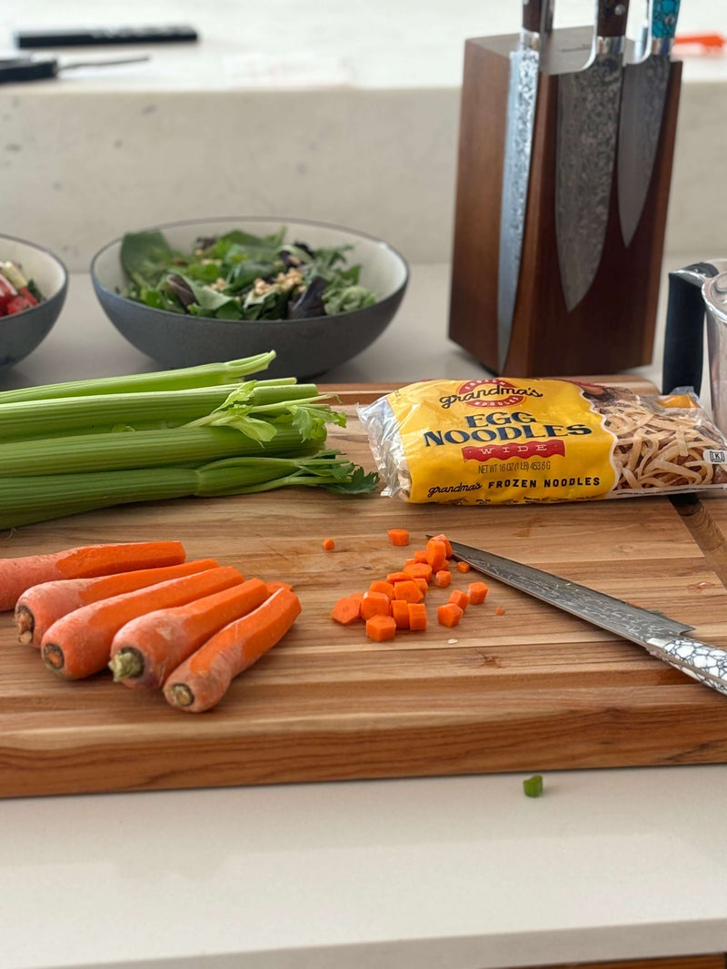 Ingredients on a cutting board for chicken noodle soup with a bag of Grandma's frozen egg noodles in the background.