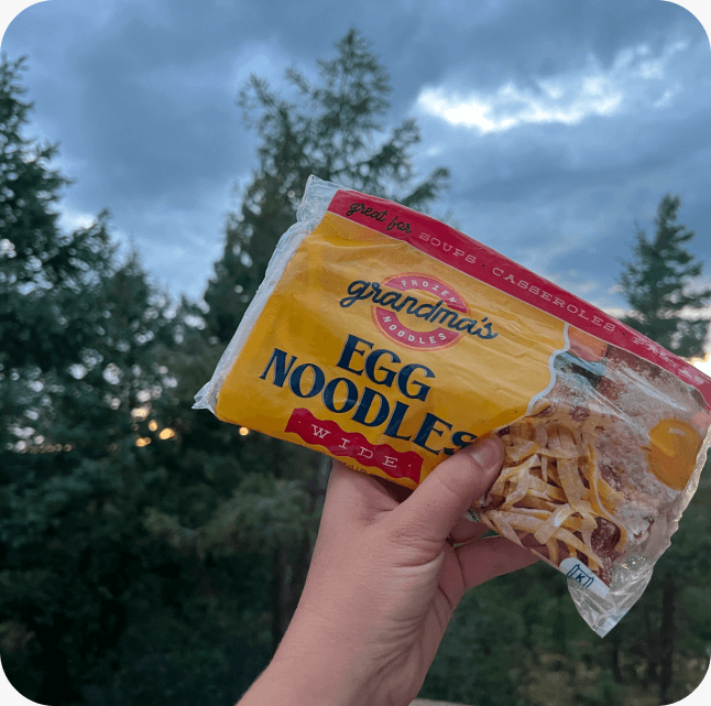 A hand holding up a package of Grandma's Frozen Egg Noodles with trees in the background